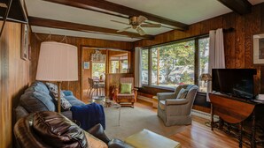 A cozy living room with wooden walls, a ceiling fan, a large window with curtains, a television, and a dining area with a table and chairs. - SUPERB VIEW BIG  SEBAGO LAKE WESTERN SIDE OF LAKE (Standish)