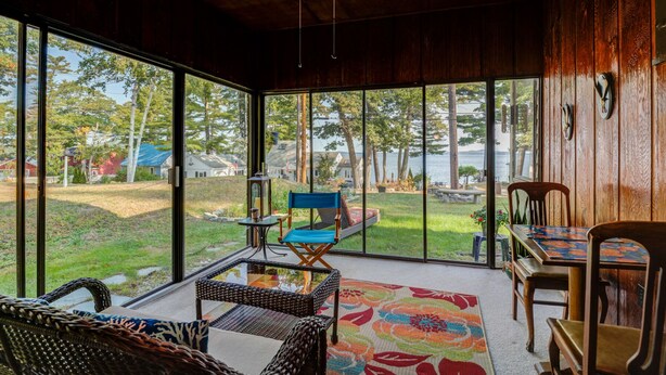 A spacious room with large windows offering a view of a lake and surrounding houses. There is a dining table with chairs, a wicker chair, and a blue cushioned chair. - SUPERB VIEW BIG  SEBAGO LAKE WESTERN SIDE OF LAKE (Standish)