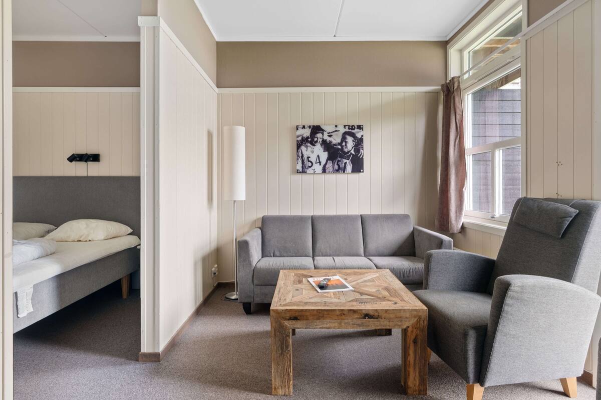 A room with a sofa, a chair, a small table, a bed, a picture on the wall, and a window.