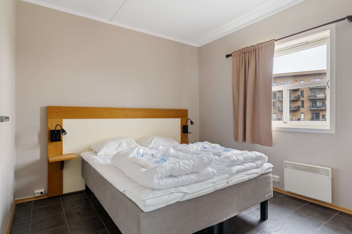 A hotel room with a bed, a window with curtains, a wall-mounted light, and a radiator.
