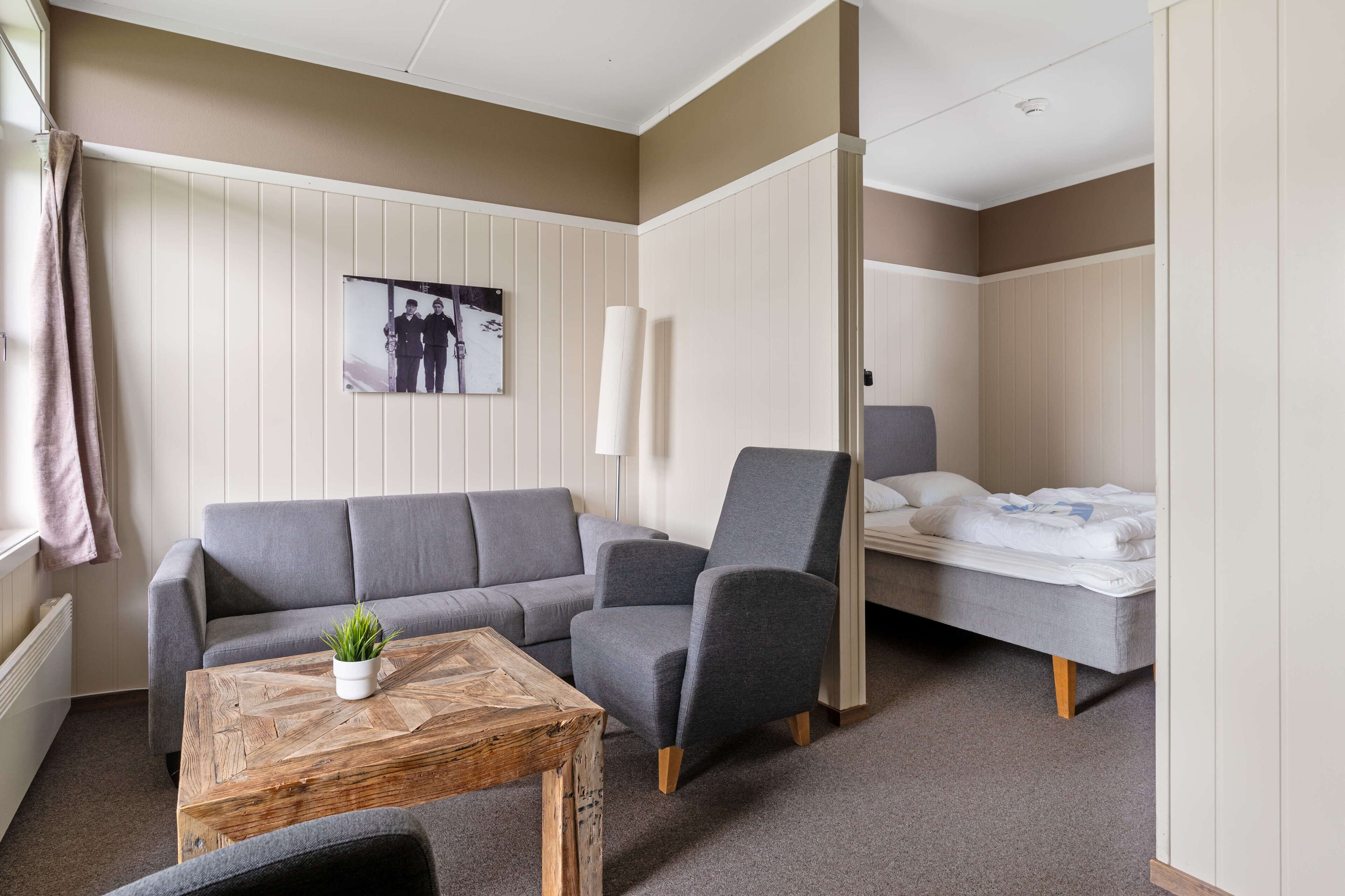 Ein Zimmer mit grauem Sofa, grauem Sessel, Couchtisch aus Holz, einem Bett und einem an der Wand hängenden Bild.