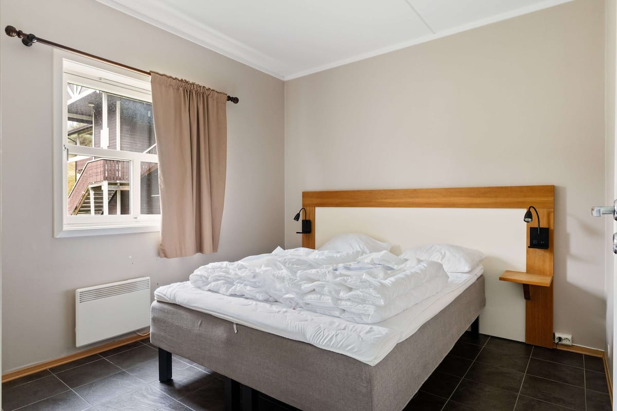 A bedroom with a bed, a window with curtains, a radiator, and a wooden headboard.