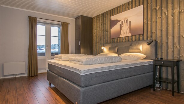 A bedroom with a large bed, wooden panel walls, a window with curtains, and a painting above the bed. - Trysilfjell Apartment Hotel (Trysil)