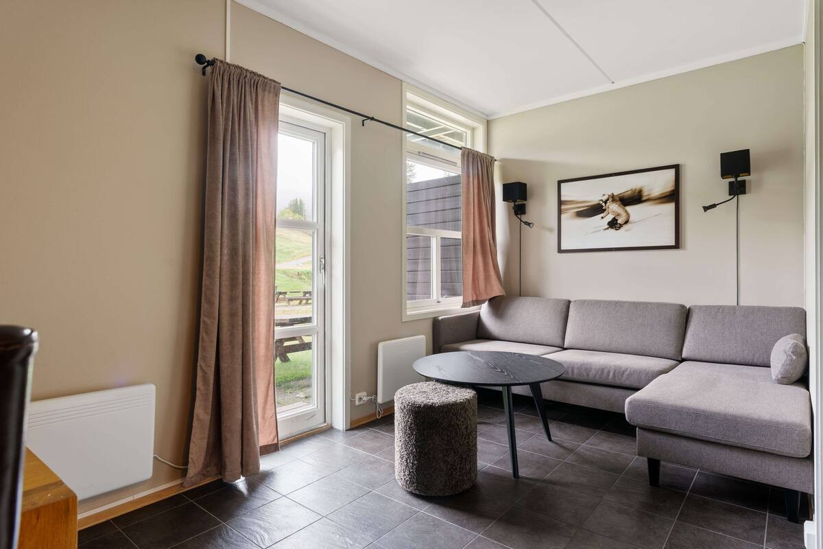 A modern living room with a grey sectional sofa, a round coffee table, a small ottoman, a radiator, and a window with curtains.