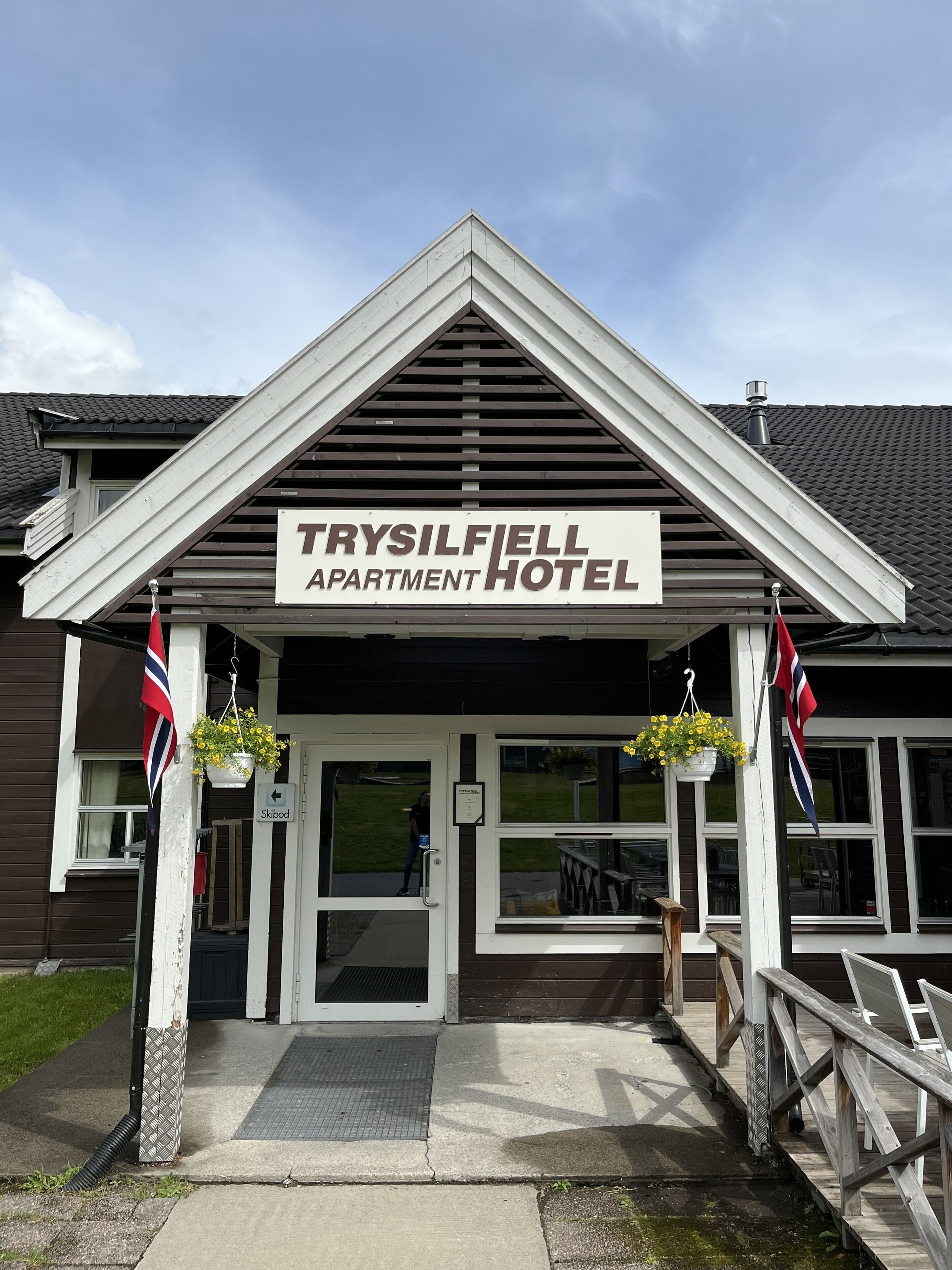 The entrance of the Trysilfiell Apartment Hotel with a clear sign, a ramp, and two flags.