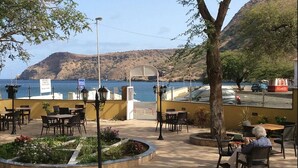 Un área de estar al aire libre con mesas y sillas, un jardín ajardinado y vistas al mar y las colinas.