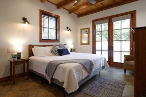 Een slaapkamer met een groot bed, houten balken, een raam met jaloezieën, een deur met uitzicht en een kleine tafel met lamp.