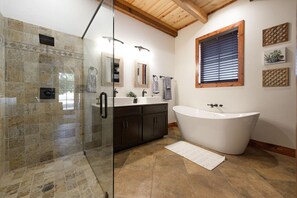 Een badkamer met een douchecabine van glas, een vrijstaand bad, donkere kastjes, een raam met jaloezieën en een kleine potplant.