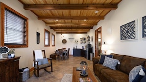 Een ruime woonkamer met een houten plafond, een bruine bank, een salontafel en een eethoek met een keuken op de achtergrond.
