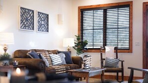 Een woonkamer met een bruine bank, twee decoratieve panelen aan de muur, een raam met jaloezieën, een bijzettafeltje met een lamp en een stoel.