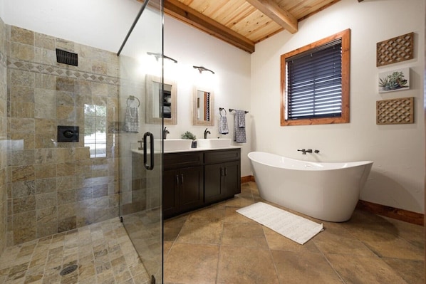 A bathroom with a glass shower enclosure, a freestanding bathtub, dark cabinetry, a window with blinds, and a small potted plant.