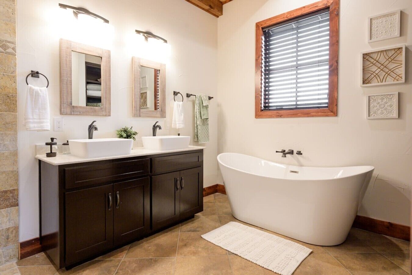 A bathroom with a freestanding bathtub, dark wooden cabinets, two sinks, and a window with blinds.