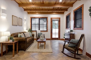 A living room with a brown leather sofa, wooden coffee table, and a rocking chair. There are two windows with blinds, a door, and a wall-mounted lamp.