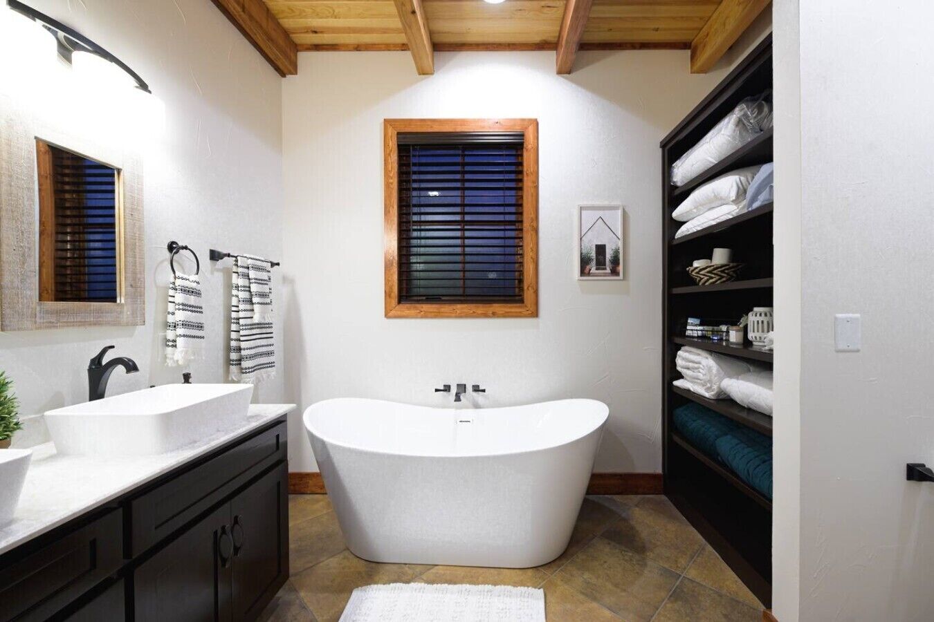 A modern bathroom with a freestanding bathtub, two sinks, a wooden ceiling, and a built-in shelf with towels.