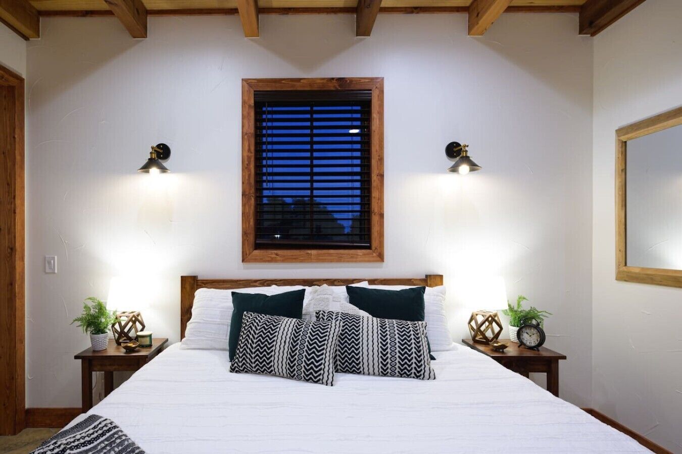 A bedroom with a wooden headboard, a large window with blinds, two bedside tables with clocks, and a bed with patterned pillows.