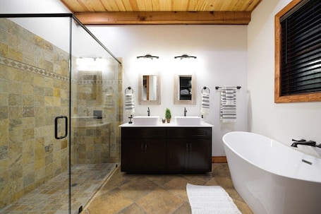 A bathroom with a glass shower enclosure, a freestanding bathtub, a double sink vanity, and a wooden ceiling.. Little Dutch Cabin #100 | Soaker Tub | Pickleball