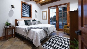 A bedroom with a bed, bedside tables, a ceiling fan, a window with blinds, a door with glass panels, and a view of the outside. - Little Dutch Cabin #100 | Soaker Tub | Pickleball (Waco)