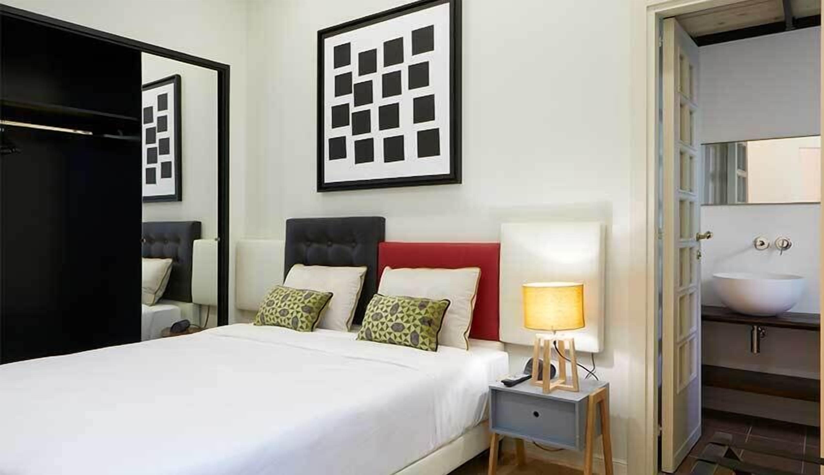 A neatly made bed with a white comforter and pillows, a bedside table with a lamp, and a large mirror above the bed.