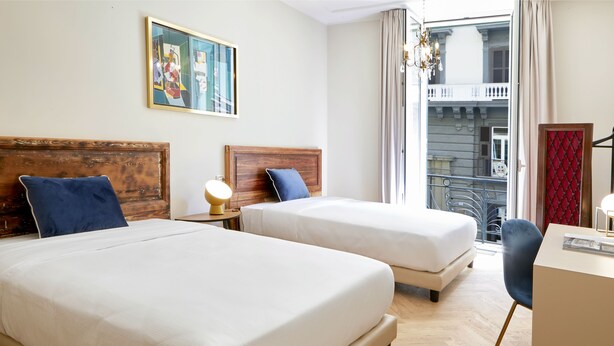 A hotel room with two beds, a desk, a chair, a painting on the wall, and a window with a view of a balcony. - Casa Vittoria Napoli - Santa Lucia (Naples)