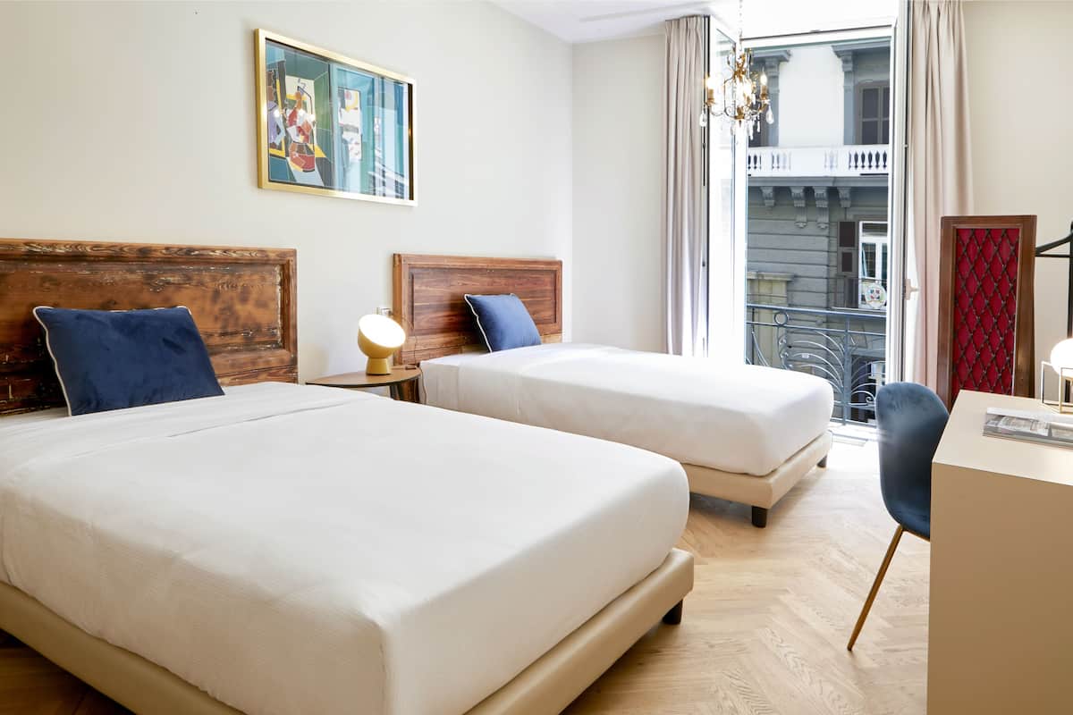 A hotel room with two beds, a desk, a chair, a painting on the wall, and a window with a view of a balcony.
