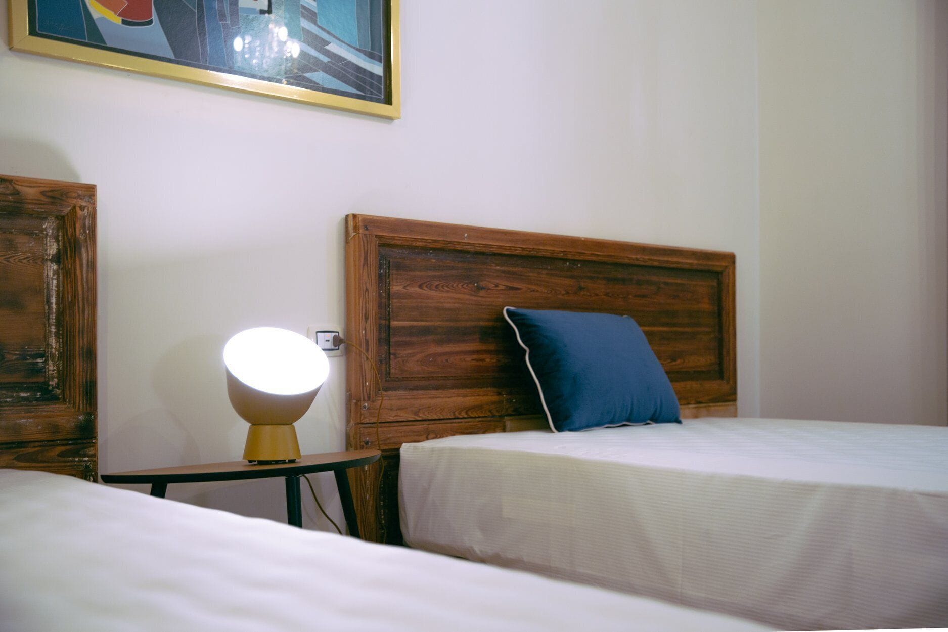 Una camera d'albergo con una testiera in legno, una lampada da comodino, un cuscino blu e un quadro appeso al muro.