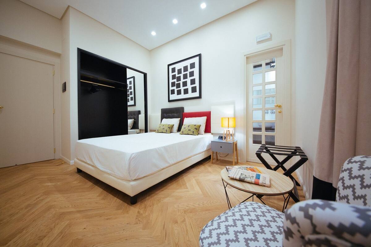 A hotel room with a bed, a nightstand, a lamp, a chair, and a small table with a newspaper.