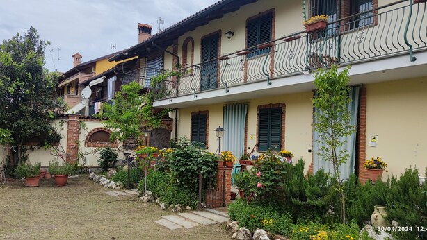 Une maison à deux étages avec une façade en briques, de nombreux balcons dotés de rampes en métal, et un jardin agrémenté de plantes et de fleurs en pot.