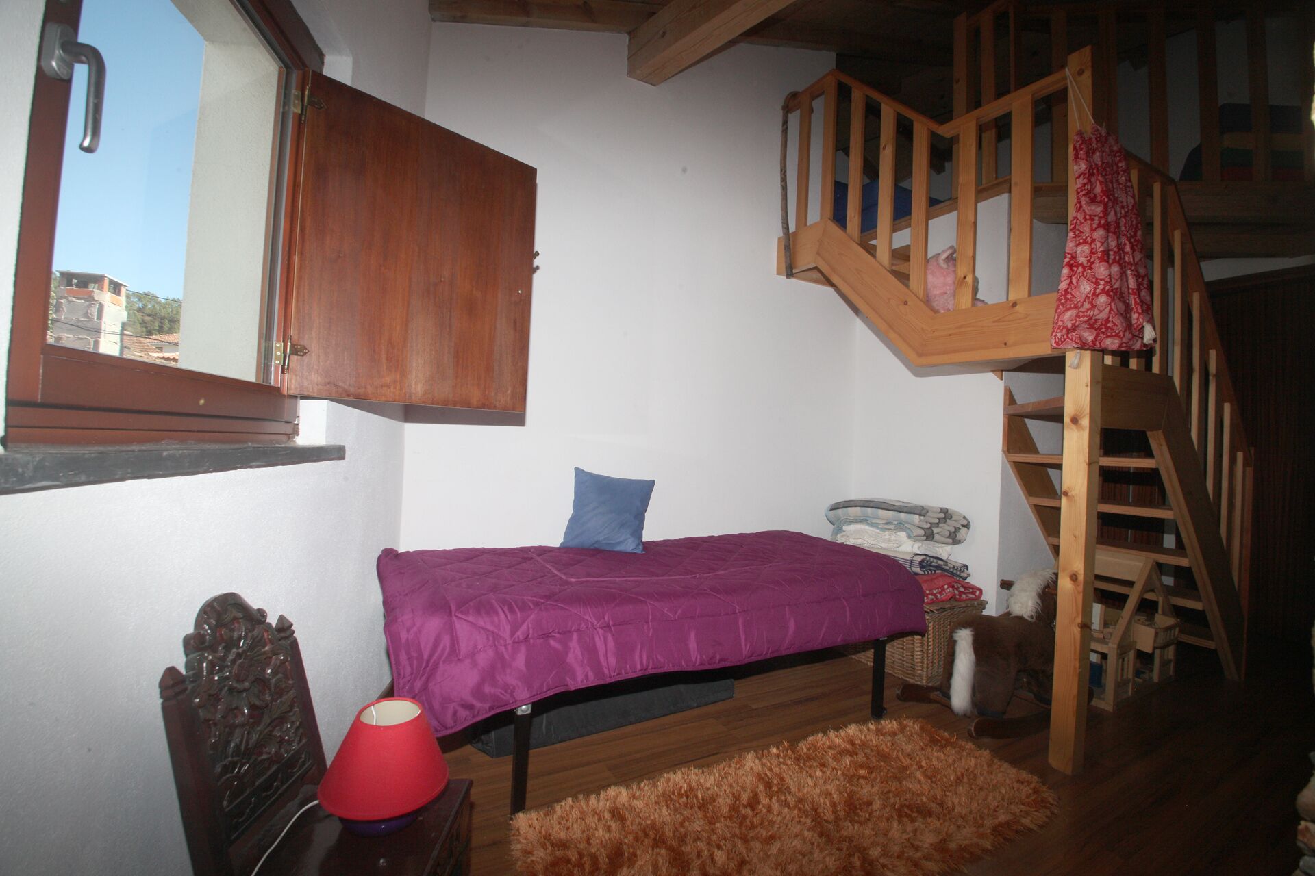 A bedroom with a wooden bunk bed, a single bed with a purple bedspread, a red lamp, a wooden headboard, a fluffy rug, and a wooden staircase.