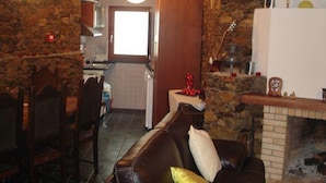 A rustic interior with a stone wall, a fireplace, a dining area with a table and chairs, and a kitchen with a window.