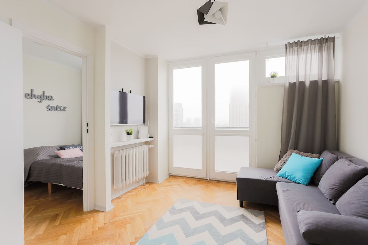 A modern living room with a sofa, a bed, a radiator, and a large window with curtains.
