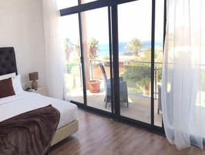 A bedroom with a large sliding glass door leading to a balcony with a view of the ocean and palm trees. - Tarrafal Guest House (Tarrafal)