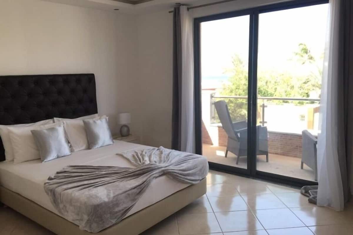 A bedroom with a large bed, a dark headboard, and a view of a balcony with outdoor furniture.
