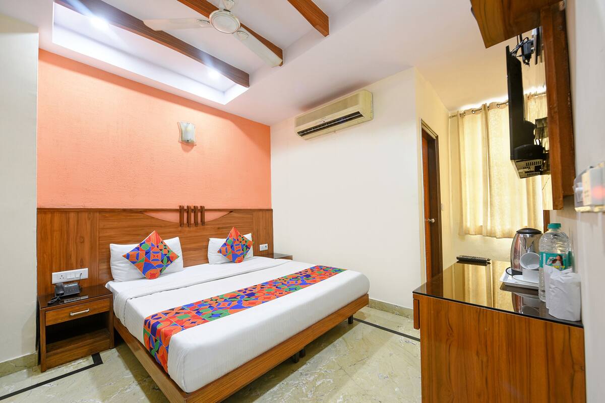 A hotel room with a wooden headboard, a bed with a colorful geometric bedspread, a nightstand with a phone, a desk with a kettle, and a window with curtains.