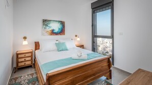 A modern bedroom with a wooden bed, a city view through a large window, and a decorative wall piece. - Morocco Mini Penthouse W Panoramic Views (Tiberias)