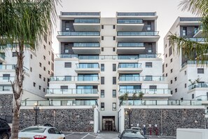 A multi-story residential building with balconies, a stone facade, and a parking area.