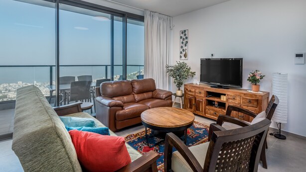A modern living room with a leather sofa, a round coffee table, and a TV on a wooden cabinet. Large windows offer a city view. - Morocco Mini Penthouse W Panoramic Views (Tiberias)