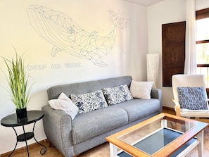 Un salon avec un canapé gris, des coussins à motifs, une table basse en verre, une œuvre d’art représentant une baleine accrochée au mur et une plante en pot.