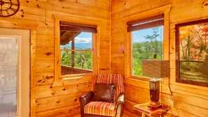 L’intérieur chaleureux d’un chalet avec un fauteuil en cuir, une table avec une lampe et de grandes fenêtres offrant une vue panoramique.