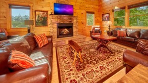 Un intérieur de chalet chaleureux avec une cheminée en pierre, des canapés en cuir, un téléviseur à écran plat et de grandes fenêtres offrant une vue sur l’extérieur.