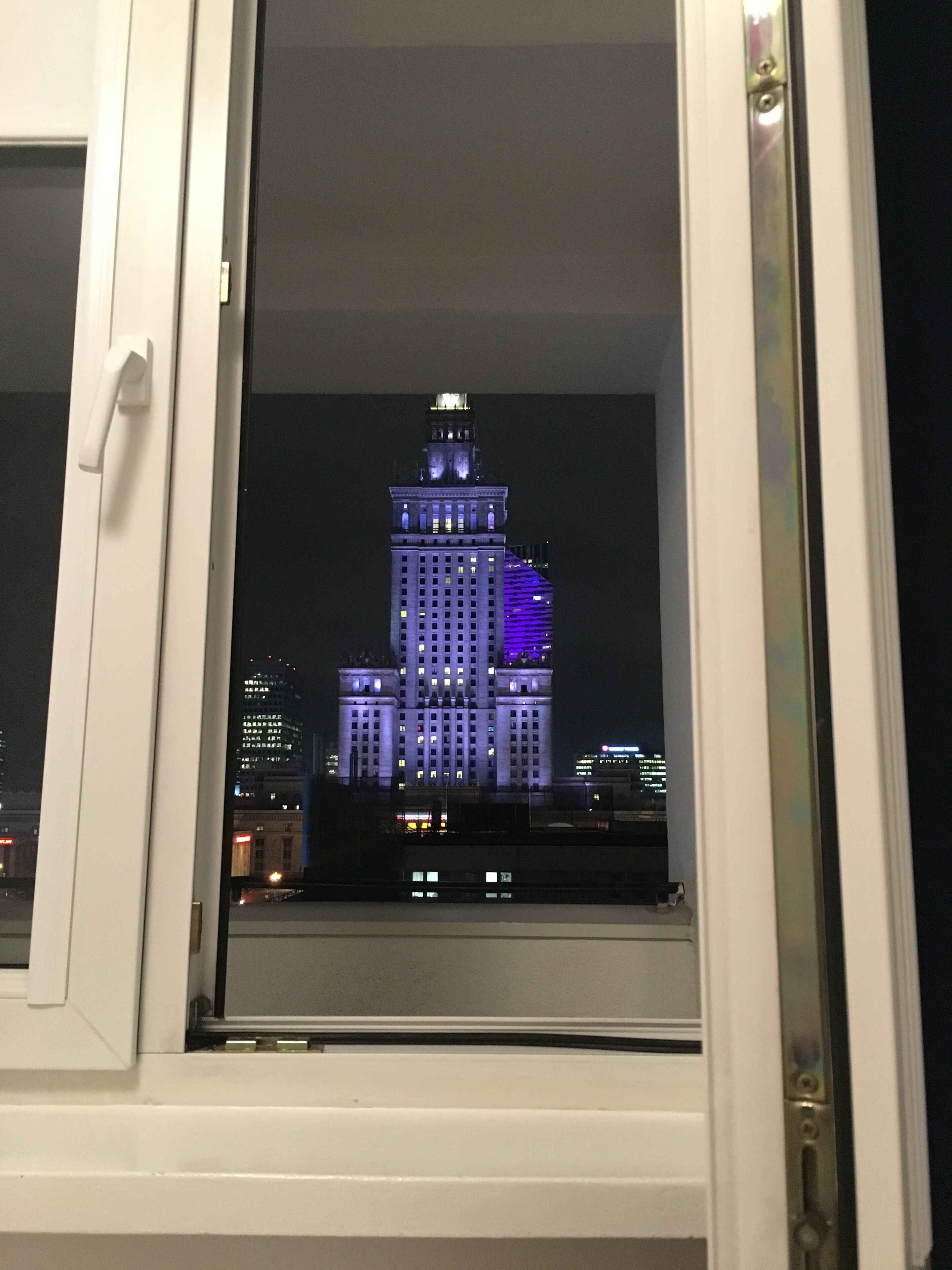 Blick auf ein Gebäude mit beleuchteten Fenstern bei Nacht, aufgenommen durch ein Fenster mit weißem Rahmen.