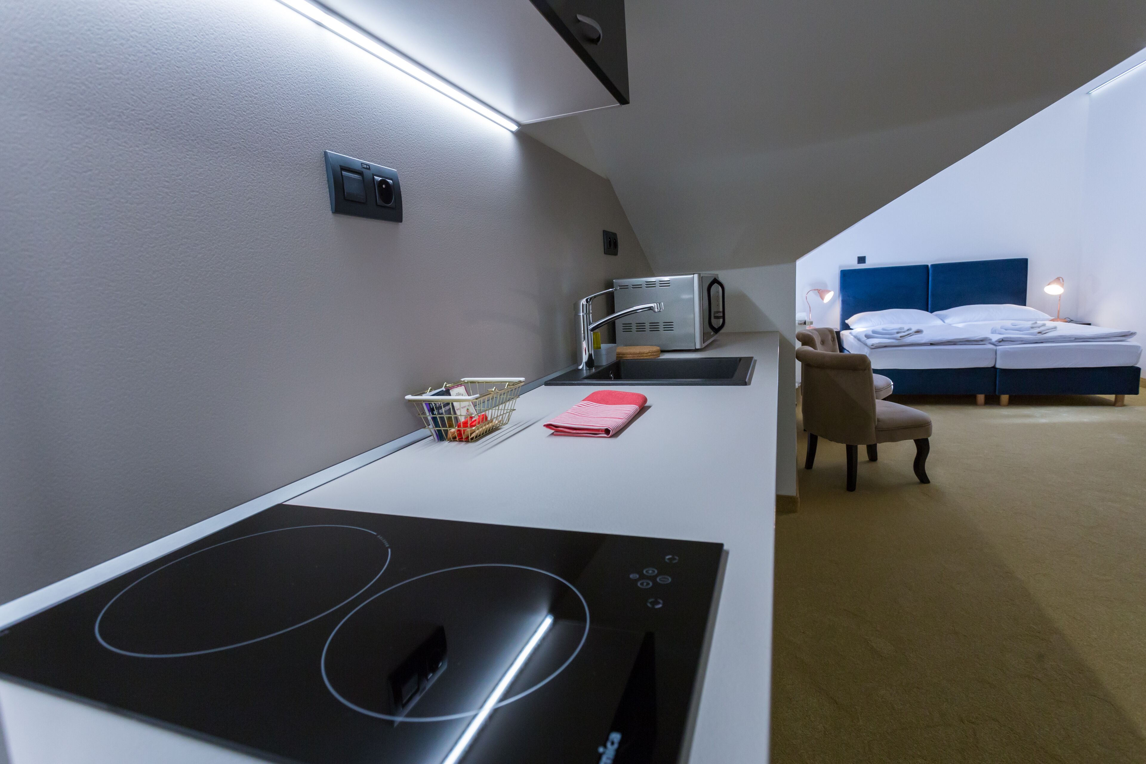 A hotel room with a kitchenette, a bed with a blue headboard, a chair, and a small table with a basket and a pink item.