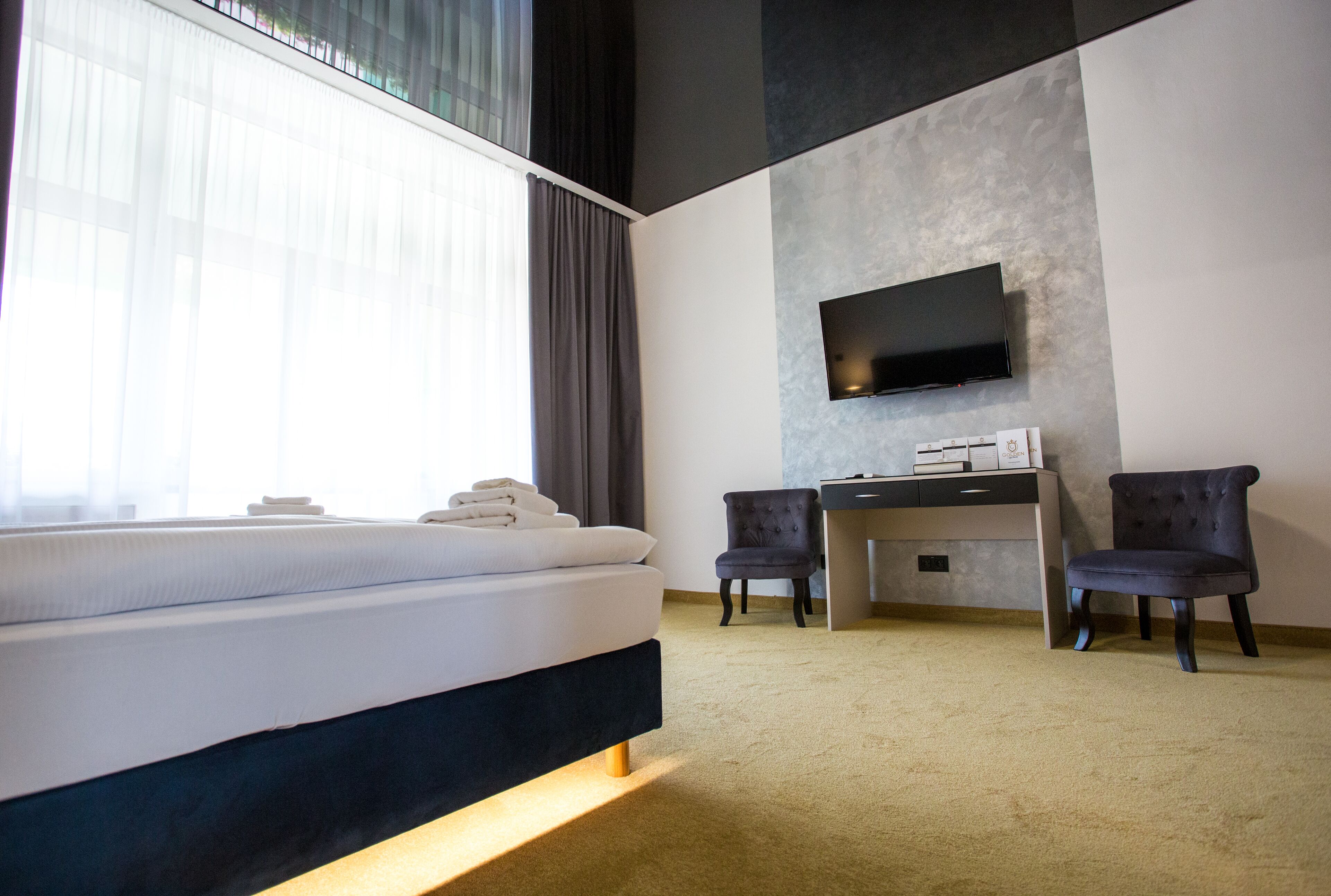 A hotel room with a bed, a desk, two chairs, a flat-screen TV mounted on the wall, and a large window with curtains.