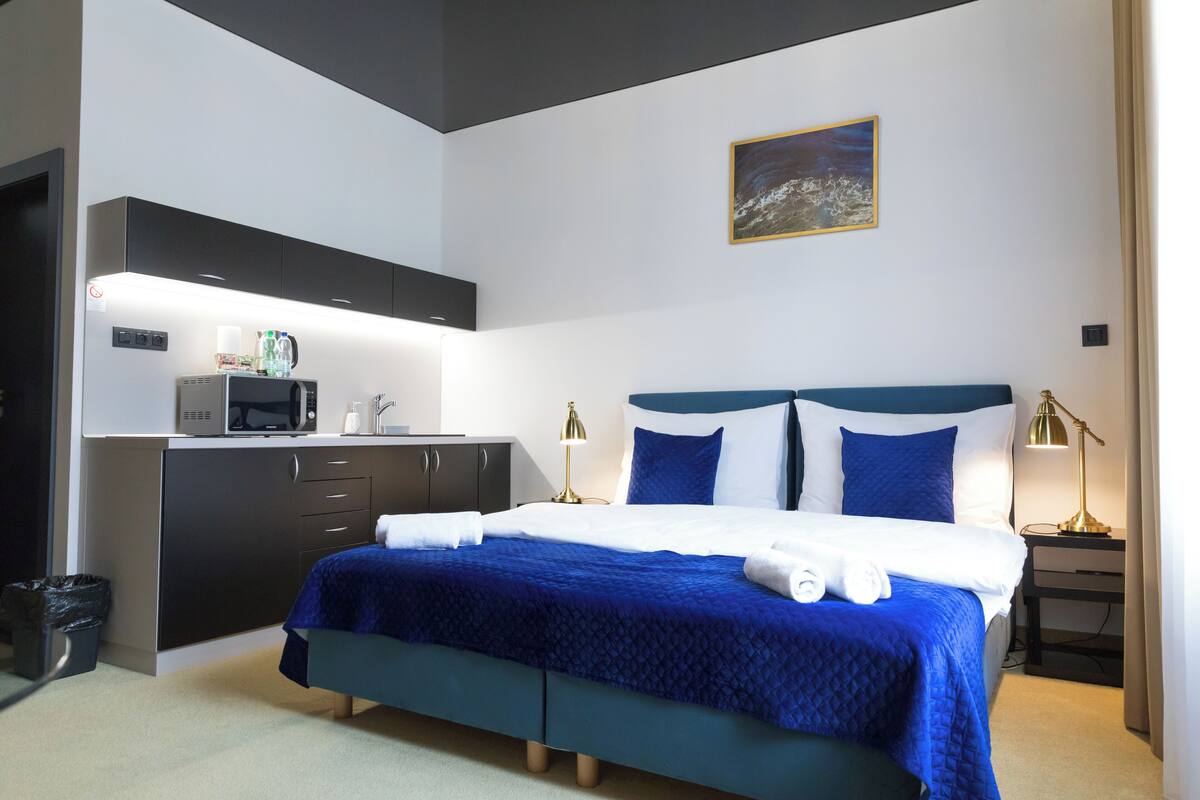 A hotel room with a bed, a desk with a microwave, a trash bin, and a painting on the wall.