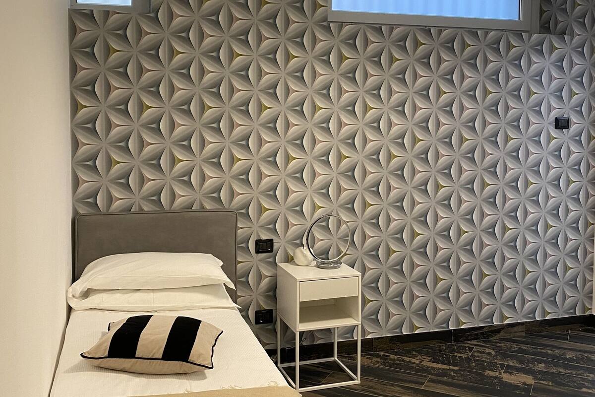 A modern bedroom with a geometric-patterned wall, a bed with white bedding, a bedside table with a mirror, and a ceiling light fixture.
