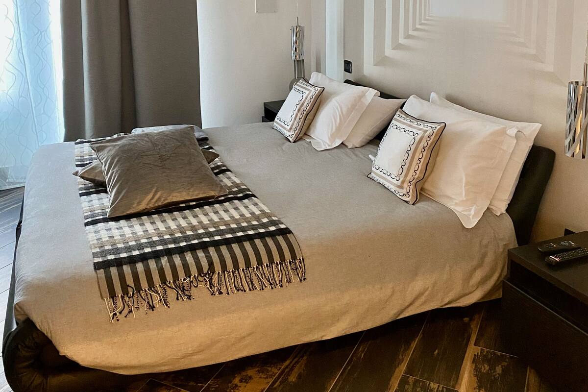 A modern bedroom with a large bed, a headboard with a geometric pattern, a bedside table with a lamp, and a window with curtains.