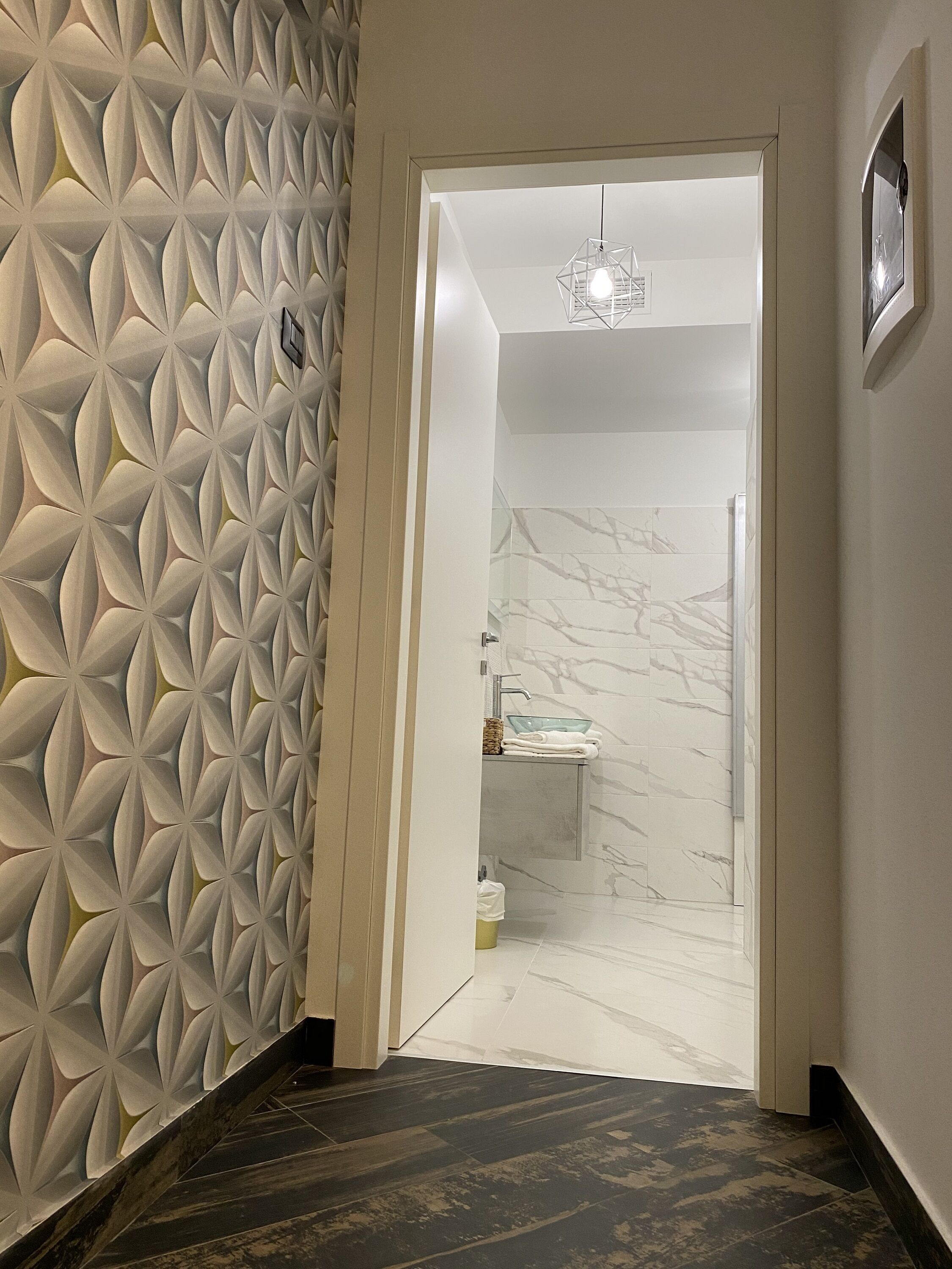 A hallway with a geometric patterned wall, a marble-tiled bathroom, and a modern light fixture.