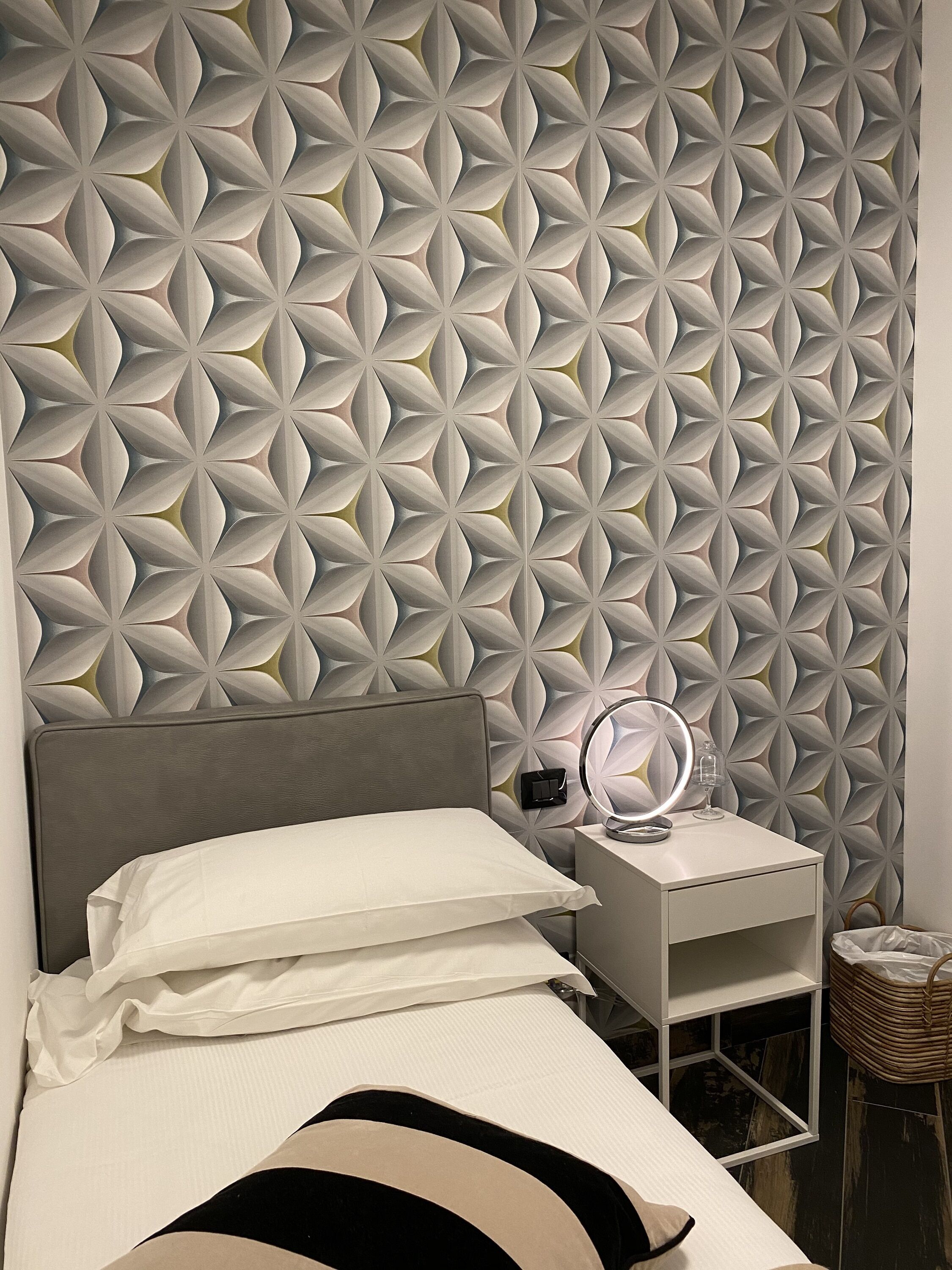 A modern bedroom with a geometric patterned wall, a bed with white bedding, a bedside table with a lamp, and a small wicker basket.