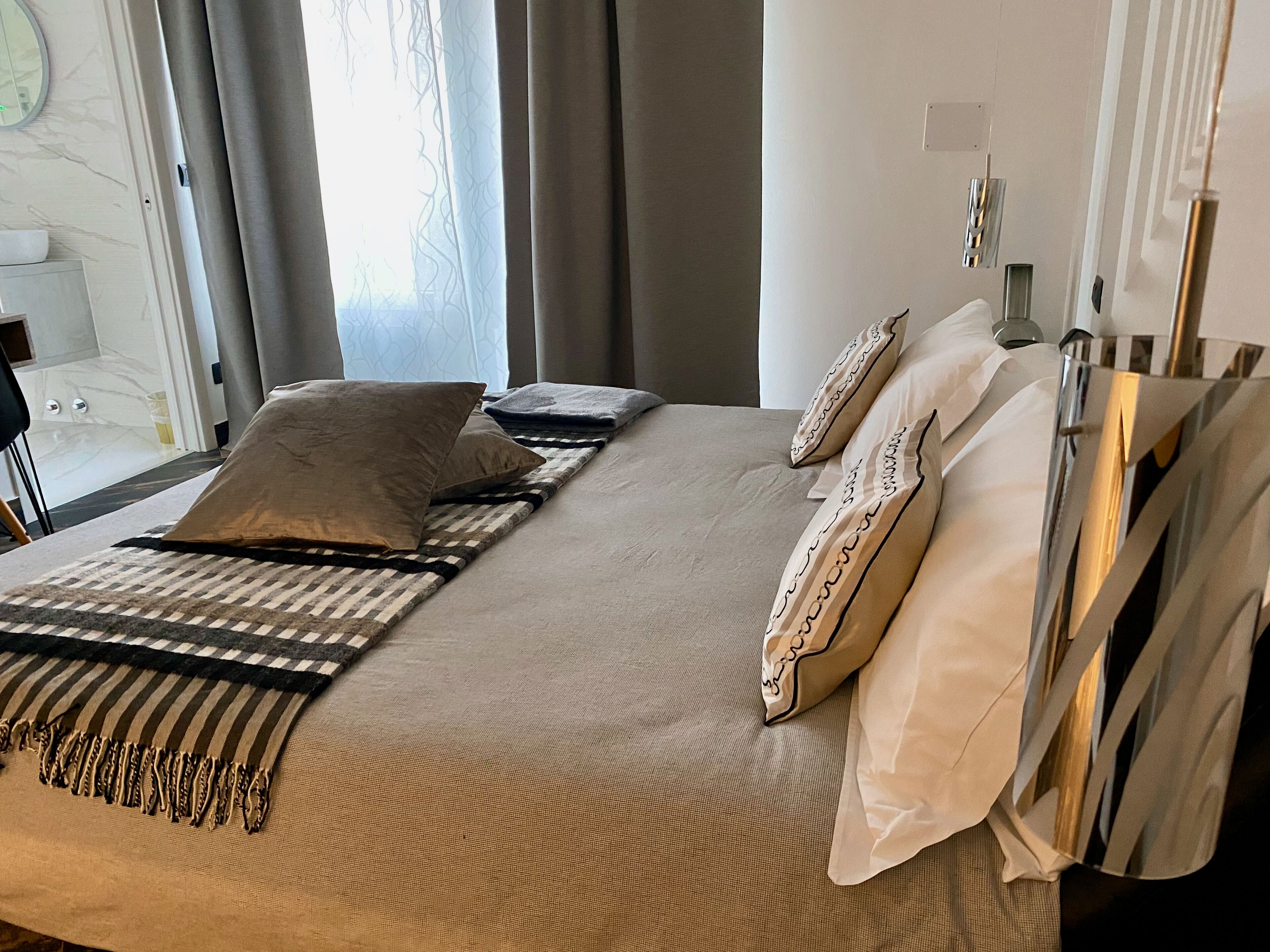 A neatly made bed with a striped blanket and pillows, a bedside table with a lamp, and a view of a bathroom with a bathtub and toilet.