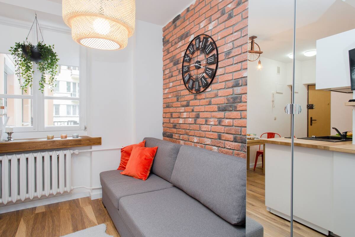 A modern living room with a grey sofa, a red cushion, a brick wall, and a large clock.