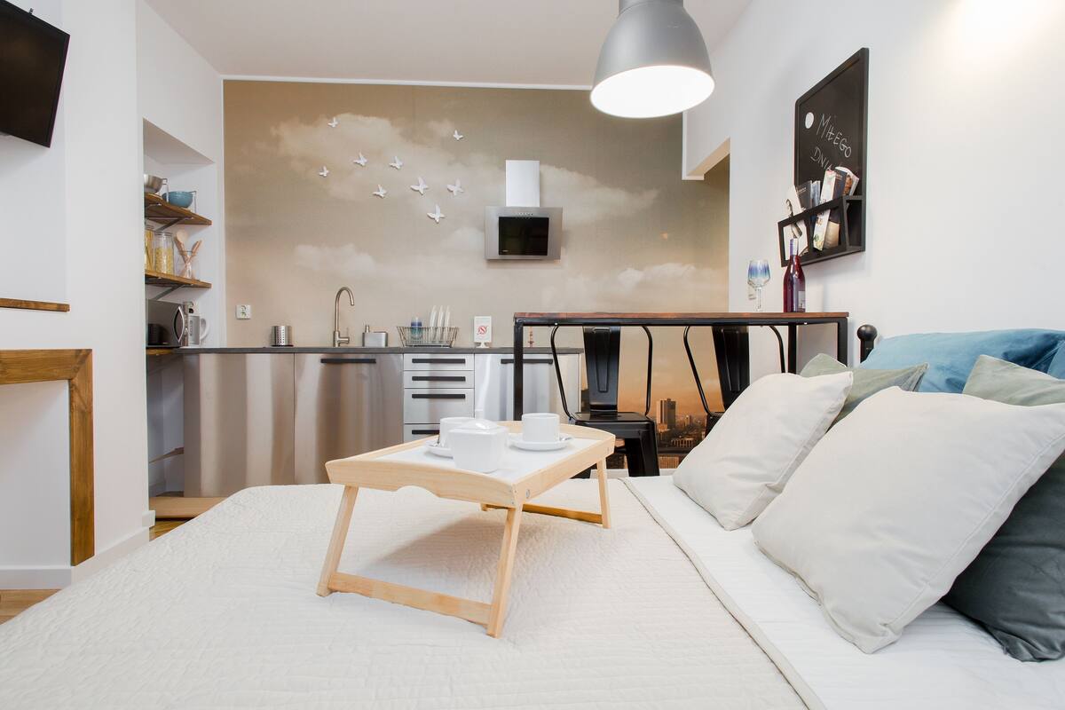 A modern bedroom with a bed, bedside table, and a view of the kitchen.
